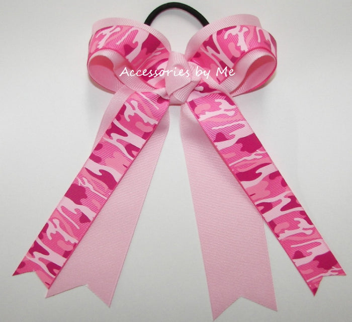 A pink hair bow accessory with a black elastic band, featuring a pink camouflage pattern on the loops and tails and a solid light pink ribbon tail, shown on a plain white background with a watermark.
