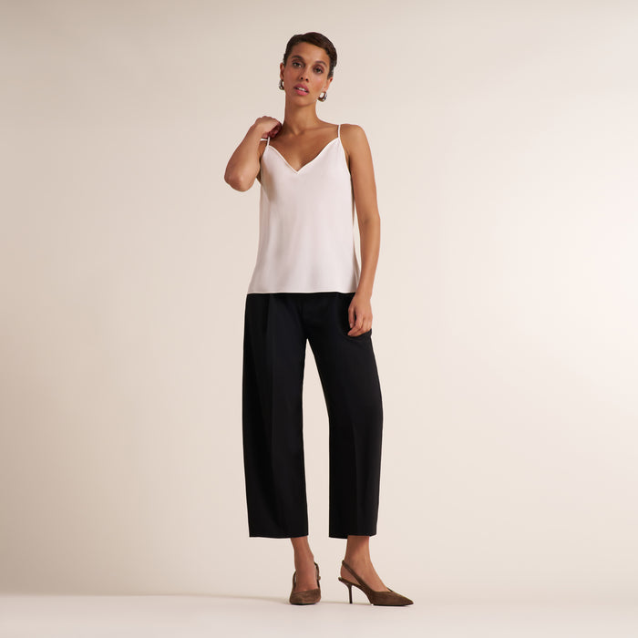 A model wearing a white spaghetti-strap camisole top and black cropped wide-leg trousers, paired with brown pointed-toe mule heels, standing against a plain beige studio background.