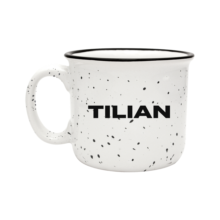 A white ceramic mug with black speckles features the Tilian brand name in bold black letters on the side, set against a solid black background.