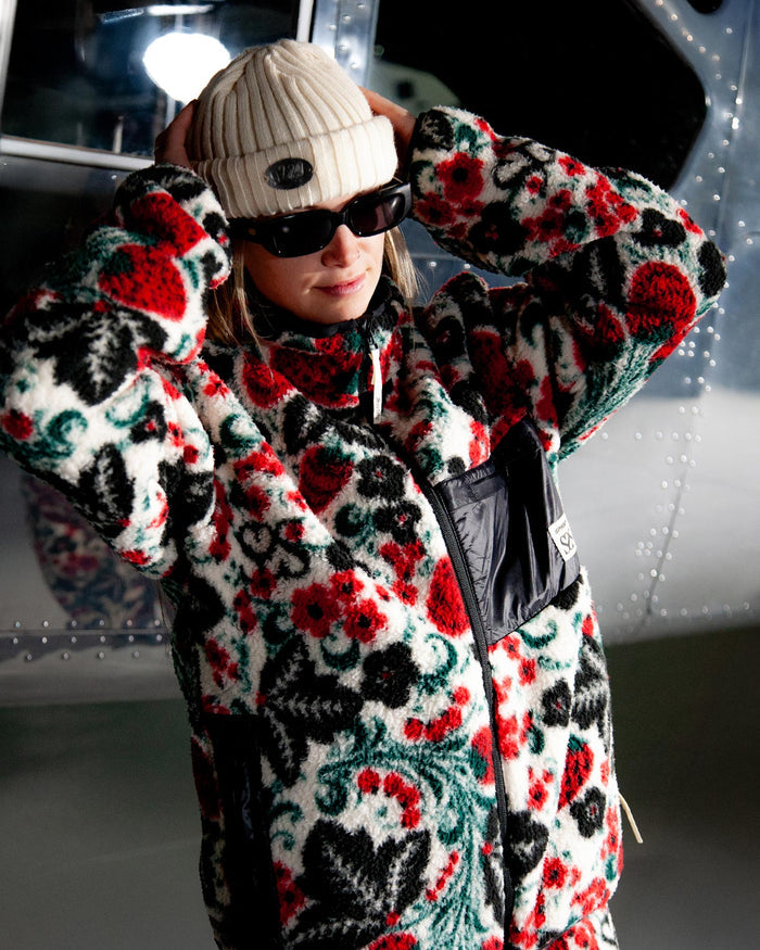 Person wearing a colorful patterned coat with a white cap and sunglasses indoors.